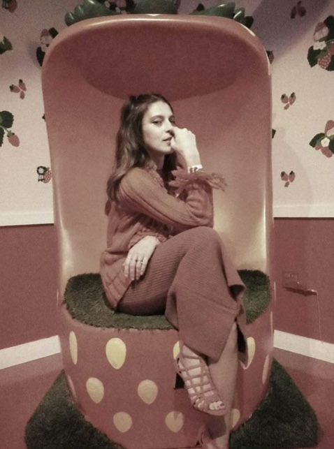 Eleanora seated sideways in a whimsical strawberry-shaped chair, glancing back over her hand