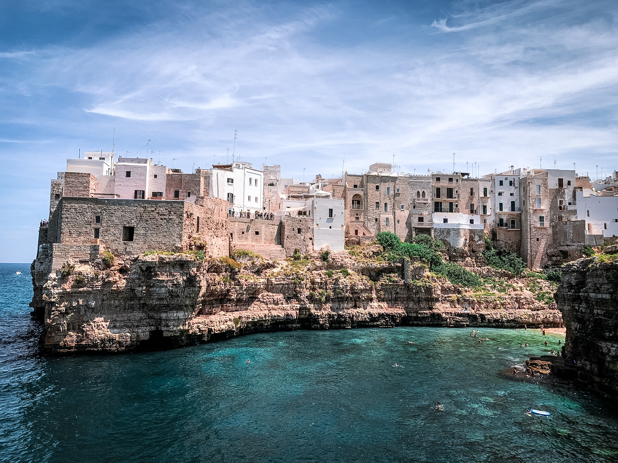 An old-world stone town perched on cliffs above a cove of clear turquoise water under a wide summer sky