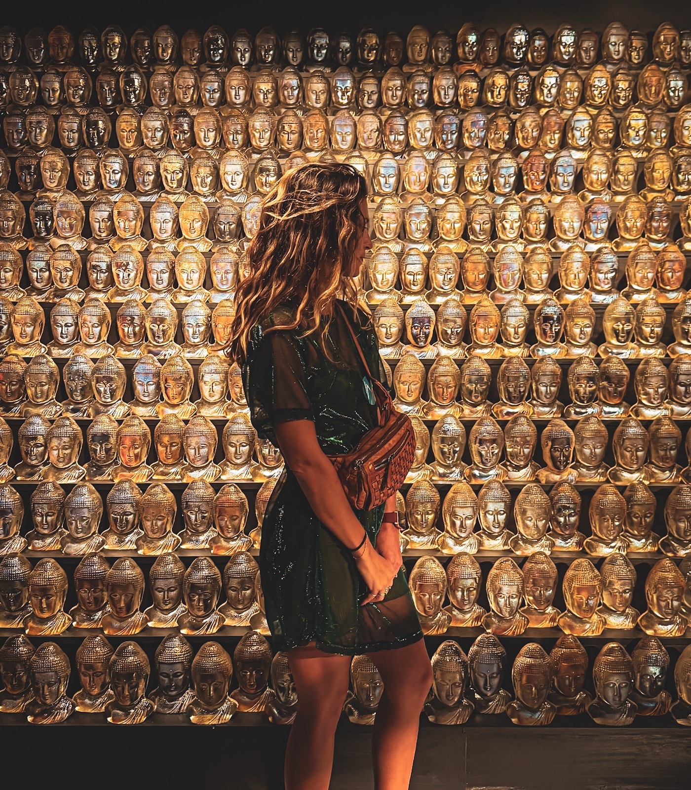 Eleanora in a sheer dark dress, in profile before a wall of hundreds of gilded golden Buddha heads lit warm